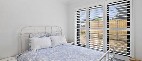 A bedroom with a bed, a bedside table, a window with blinds, and a view of a fence.