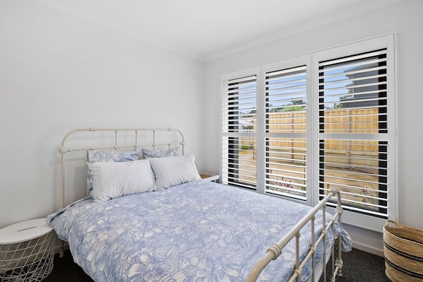 A bedroom with a bed, a bedside table, a window with blinds, and a view of a fence.
