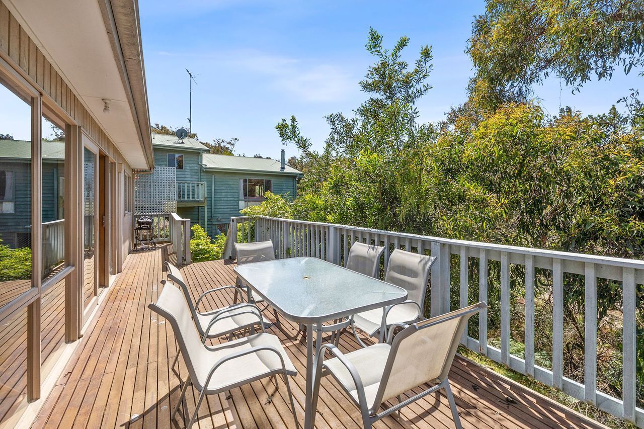 Une terrasse en bois, agrémentée d’une table et de chaises, entourée d’une balustrade blanche, et offrant une vue sur un jardin et une maison.