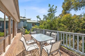 Eine Holzterrasse mit Tisch und Stühlen, umgeben von einem weißen Geländer, mit Blick auf einen Garten und ein Haus.