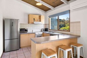 Eine moderne Küche mit Edelstahl-Kühlschrank, Mikrowelle und Herd, Holzschränken, einer großen Kochinsel mit Hockern und einem Fenster mit Blick auf Grünflächen.
