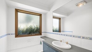 A bathroom with a bathtub, a sink, and a window with a view of greenery. - Oasis on Weir (Anglesea)