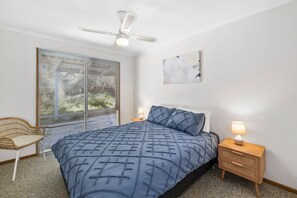A bedroom with a bed, a chair, a bedside table with a lamp, a ceiling fan, and a window with blinds. - Oasis on Weir (Anglesea)