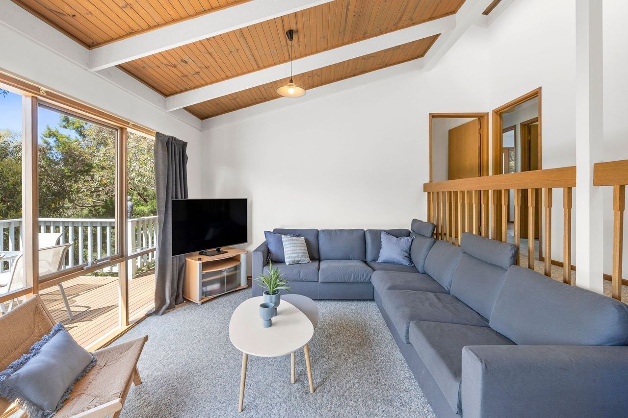 A modern living room with a sectional sofa, a flat-screen TV, a small round table, and a balcony with outdoor seating.