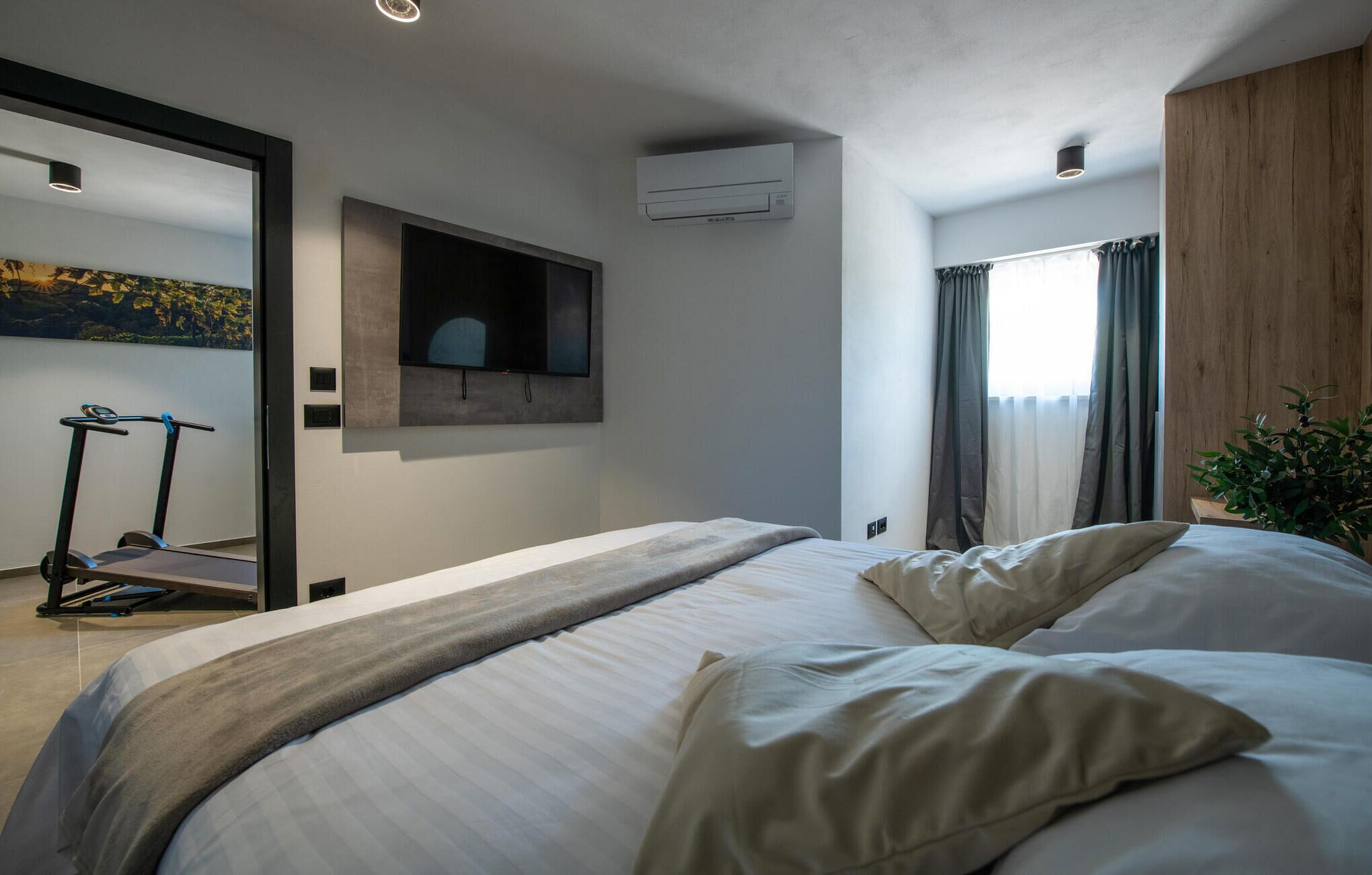 Una moderna camera d'albergo con un letto, un tapis roulant, un condizionatore e una grande finestra con tende.