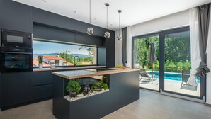A modern kitchen with a central island, built-in oven, and a view of a swimming pool and greenery. - Gorgeous home in Nedescina with sauna (Nedescina)