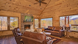 A log cabin interior with a stone fireplace, leather furniture, and large windows offering views of the forest. - 🏔️A Wild Mountain Splash: Mtn View, Pets, Secluded, Theater, Games, Hot Tub (Sevierville)
