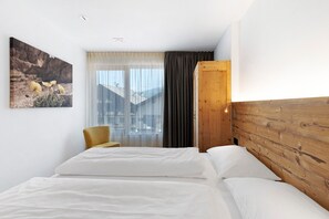 A hotel room with a bed, a chair, a wooden headboard, a window with curtains, and a painting on the wall.