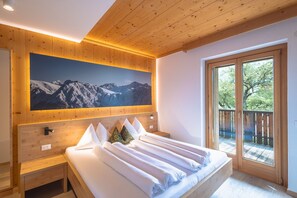 A bedroom with a wooden ceiling, a large bed with white linens, a nightstand, a wooden headboard, a painting of mountains, and a balcony with a view of trees.