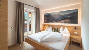 Une chambre à coucher comprenant un lit, une table de chevet, une lampe fixée au mur, une fenêtre avec des rideaux et un grand tableau représentant un paysage.