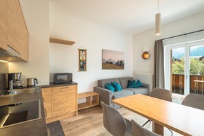Een moderne keuken met een inbouwoven, een eethoek met een houten tafel, een grijze bank met blauwe kussens en uitzicht op de omgeving door een groot raam.