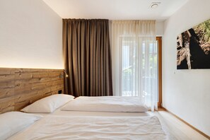 A bedroom with a wooden headboard, a bed with white linens, a window with sheer curtains, and a wall-mounted picture.