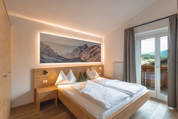 Une chambre avec un grand lit, une table de chevet, un banc en bois, un tableau représentant des montagnes et une vue à travers une porte vitrée.