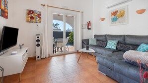A living room with a grey sofa, a flat-screen TV, a white TV stand, and a sliding glass door leading to an outdoor area.
