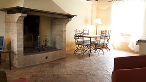 A cozy living room with a stone fireplace, wooden ceiling beams, a dining table with chairs, and a red sofa. - Benozzo vacation apartment with garden and pool (Montefalco)