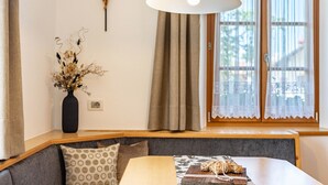 Una habitación con un banco de madera, una mesa con una cesta tejida, un jarrón con flores secas y una cruz en la pared.