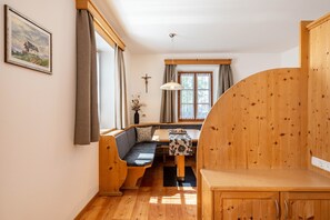 Une pièce chaleureuse avec un banc en bois, une table recouverte d’une nappe à motifs floraux, un crucifix au mur et une fenêtre avec des rideaux.