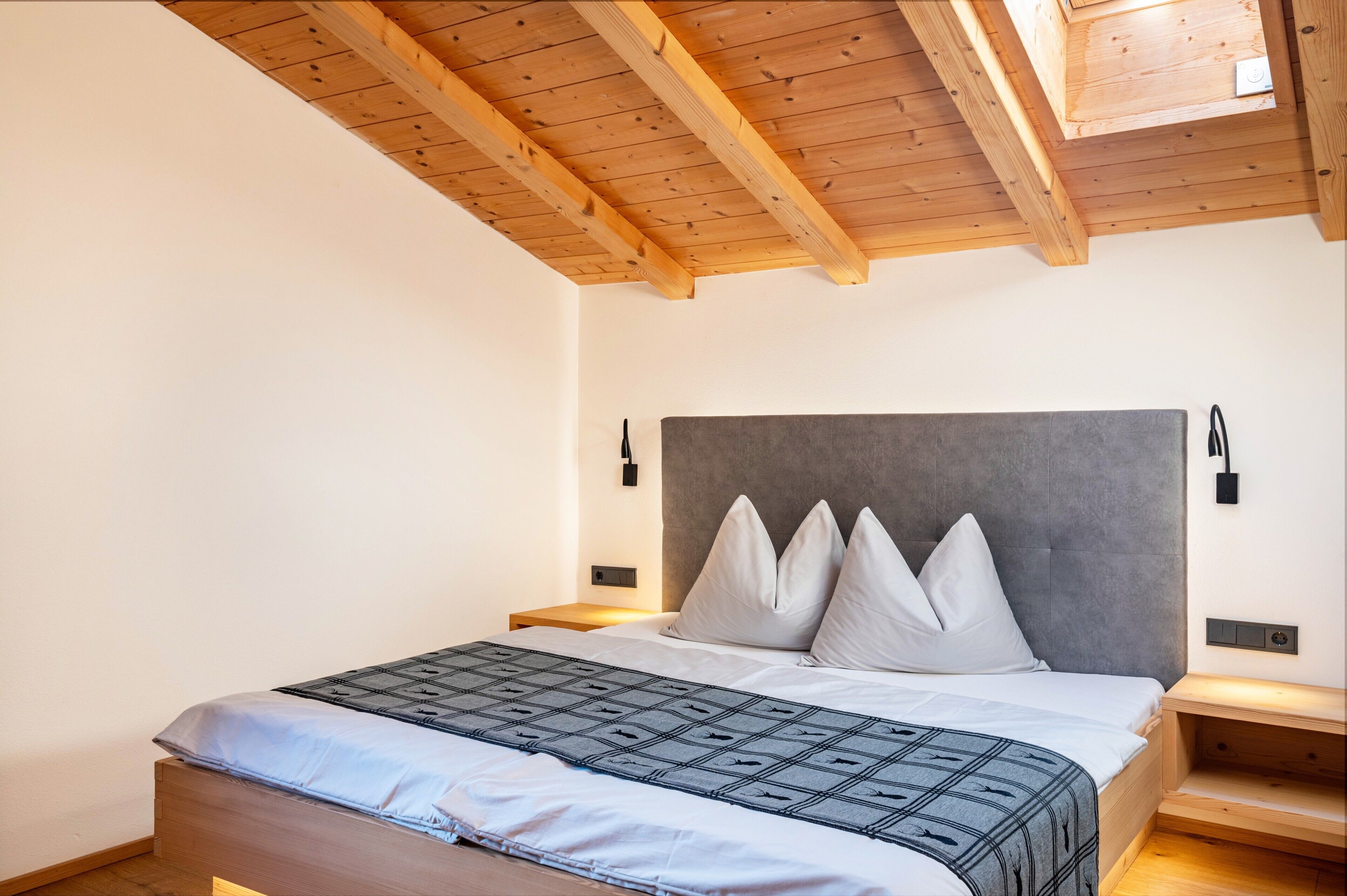 Een slaapkamer met een houten plafond, een bed met een grijs hoofdeinde, witte kussens en een grijze gebloemde deken.