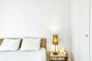 Cama com roupa de cama branca, cabeceira de madeira, mesa de cabeceira com abajur e velas.