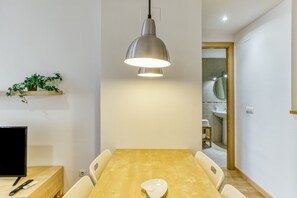 Uma sala de jantar com mesa de madeira, cadeiras brancas, luminária pendente, televisão e planta em uma estante.
