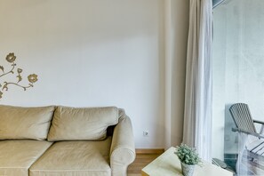Um sofá bege, uma mesinha com uma planta e uma cadeira perto de uma janela com cortinas.
