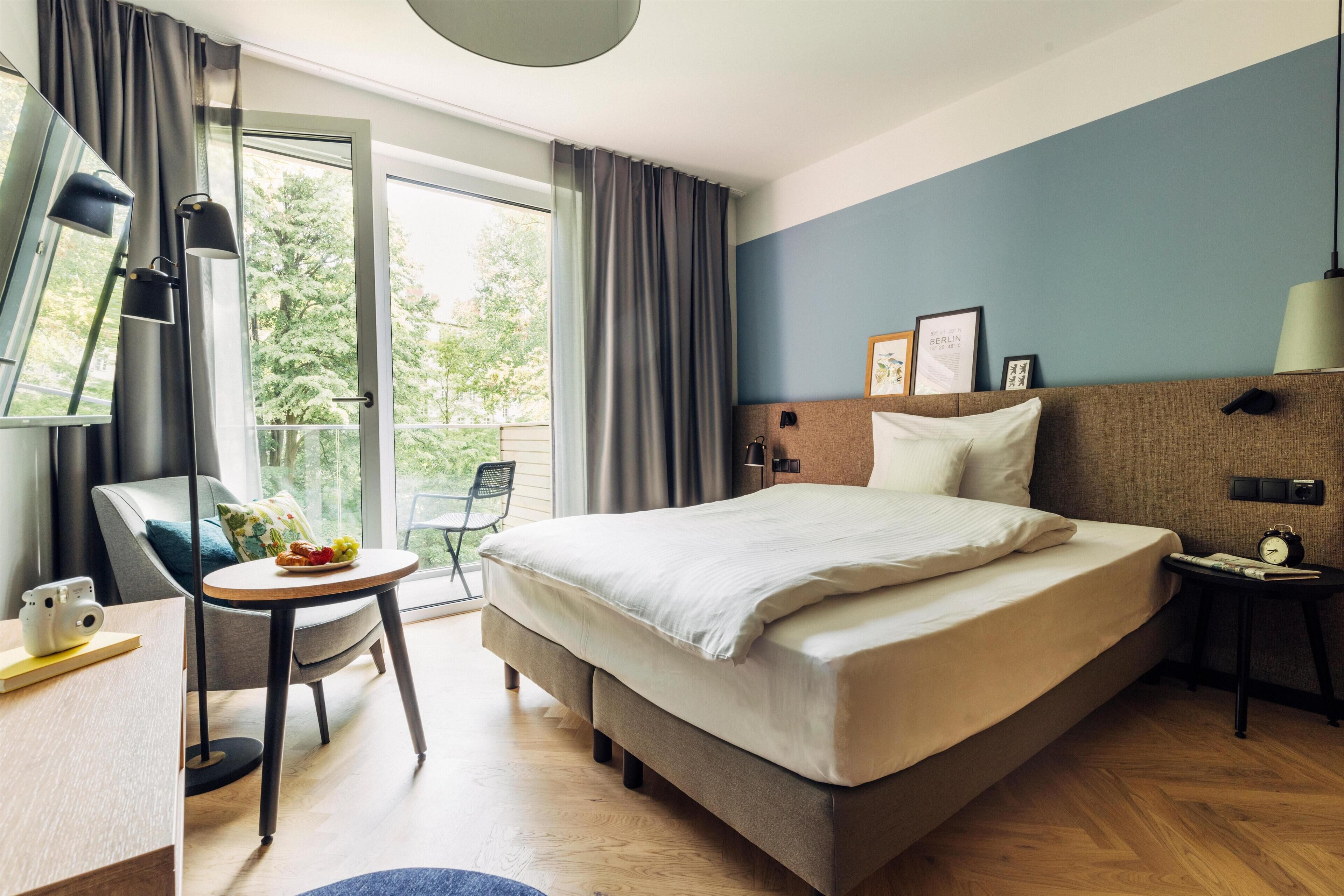 A modern hotel room with a large bed, a desk with a lamp, a chair, and a view of the outdoors.