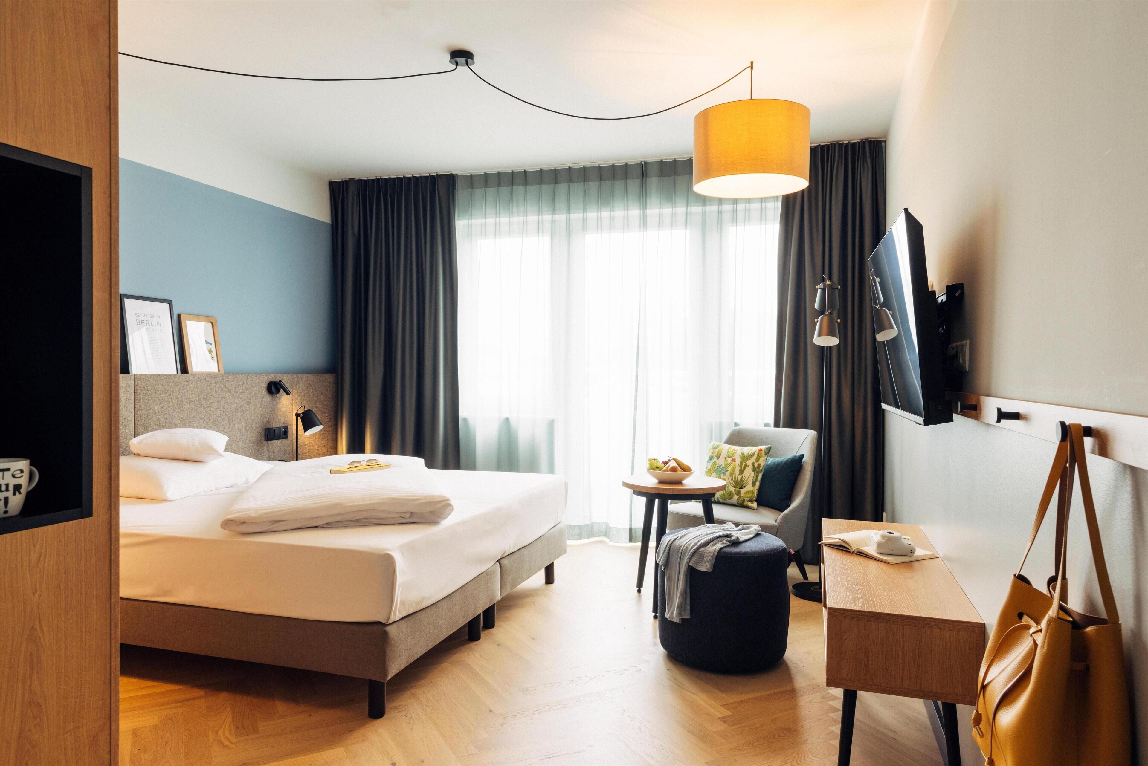 A hotel room with a bed, a desk, a chair, a TV, and a window with curtains.