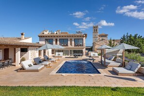 A pool area with lounge chairs and umbrellas, a clear blue sky, and a building with a tower in the background. - Vacation Home 'Sa Figuereta Living Place' with Mountain Views, Wi-Fi & Air Conditioning (ARIANY)