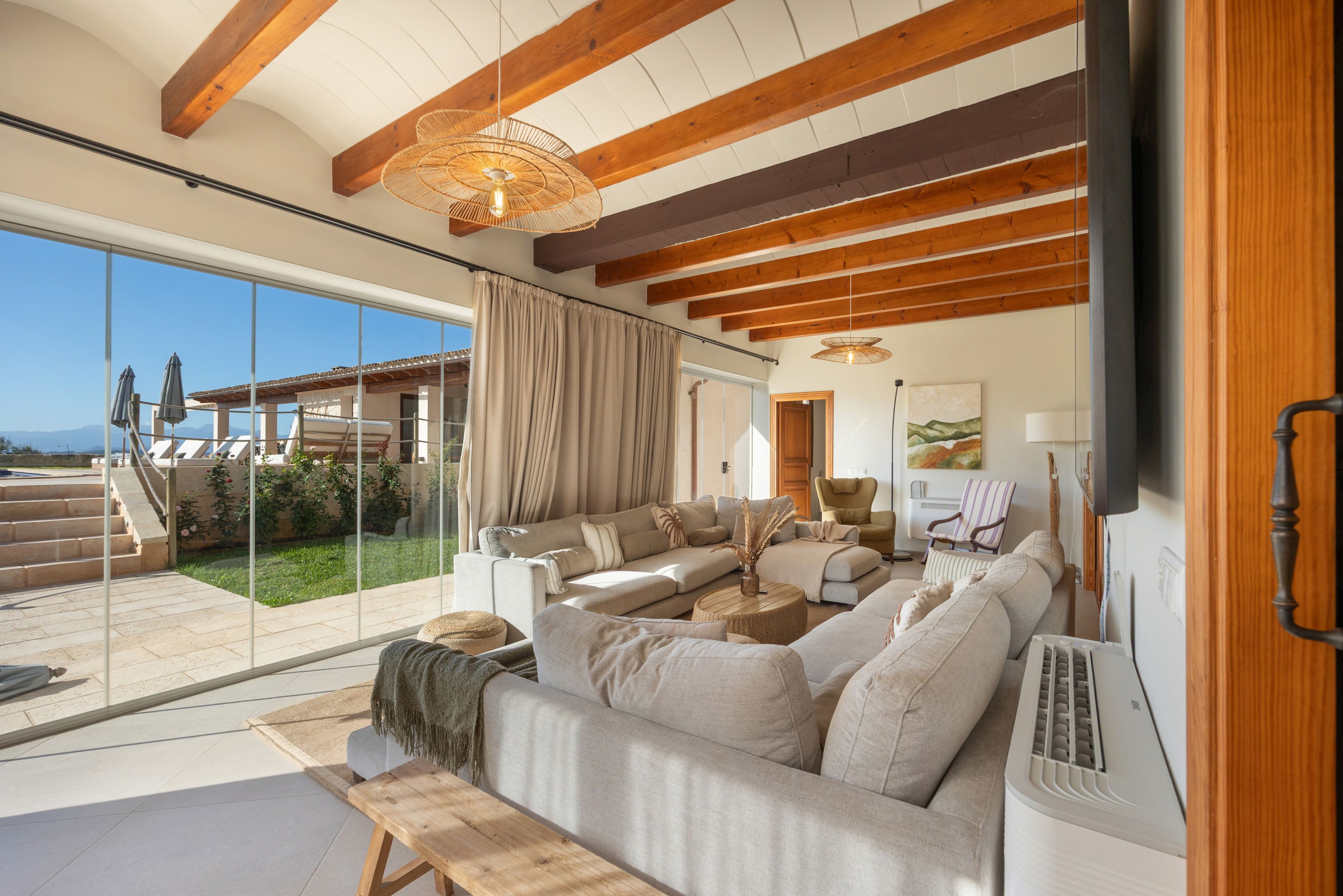 Amplia sala de estar con un sofá grande, un banco de madera y una mesa de comedor. Hay una puerta de cristal que da a un patio exterior con vistas a las montañas.