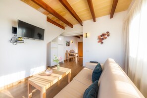 Un salon moderne doté d’un téléviseur à écran plat, de poutres en bois, d’un canapé, d’un coin repas et d’une cuisine.