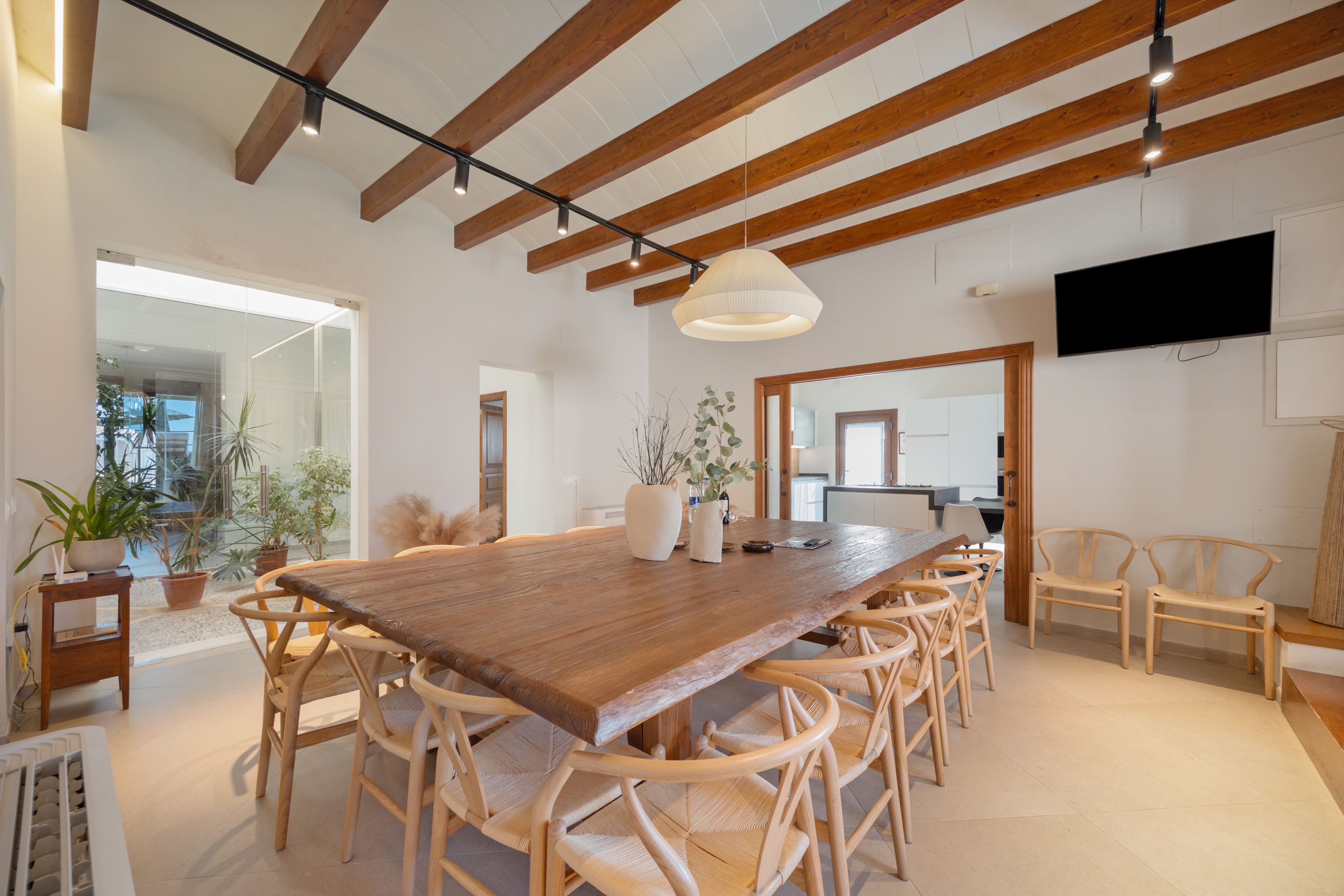 A spacious dining area with a long wooden table surrounded by wicker chairs, a hanging light fixture, and a television mounted on the wall.