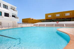 Una piscina con agua azul cristalina, rodeada por un edificio blanco con balcones y un edificio amarillo al fondo.