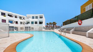 Piscina com borda curva, rodeada por um edifício branco com varandas, um flutuador salva-vidas e um céu azul límpido.