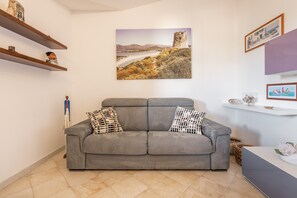 A living room with a grey sofa, a painting of a coastal landscape, and shelves with decorative items.