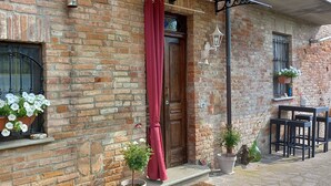 A brick building with a wooden door, a window with white flowers, a red curtain, and outdoor seating.