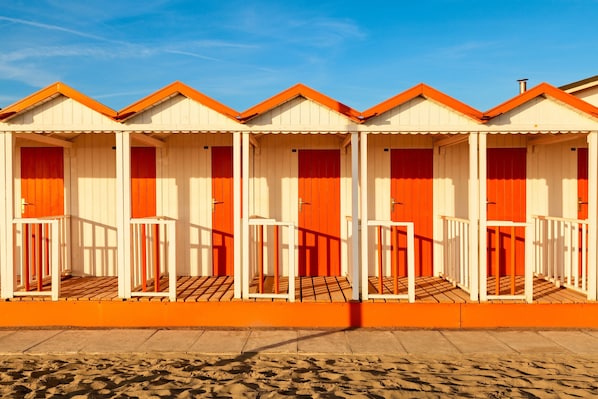 Une rangée de cabanes de plage aux façades blanches et orange, avec des planchers en bois, sous un ciel d’un bleu limpide.