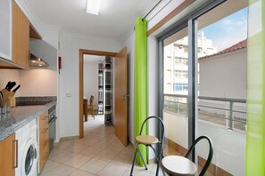 A kitchen with a washing machine, a dining area with a chair, and a balcony with a view of buildings.