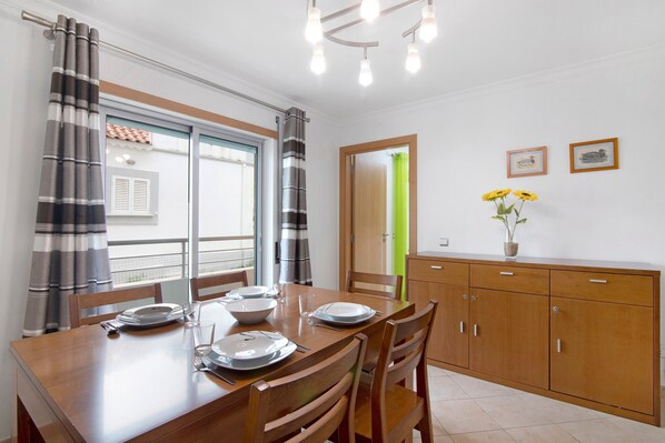 A dining area with a wooden table set for four, chairs, a cabinet with drawers, and a window with curtains.