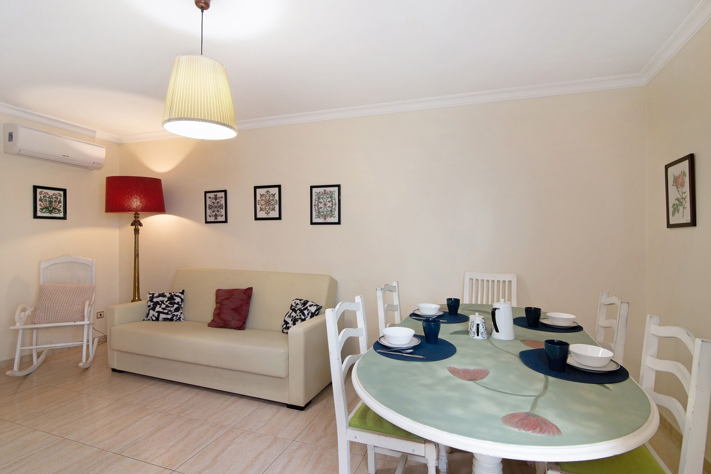 Een eethoek met een ronde tafel, witte stoelen, een bank, een schommelstoel en een hanglamp.