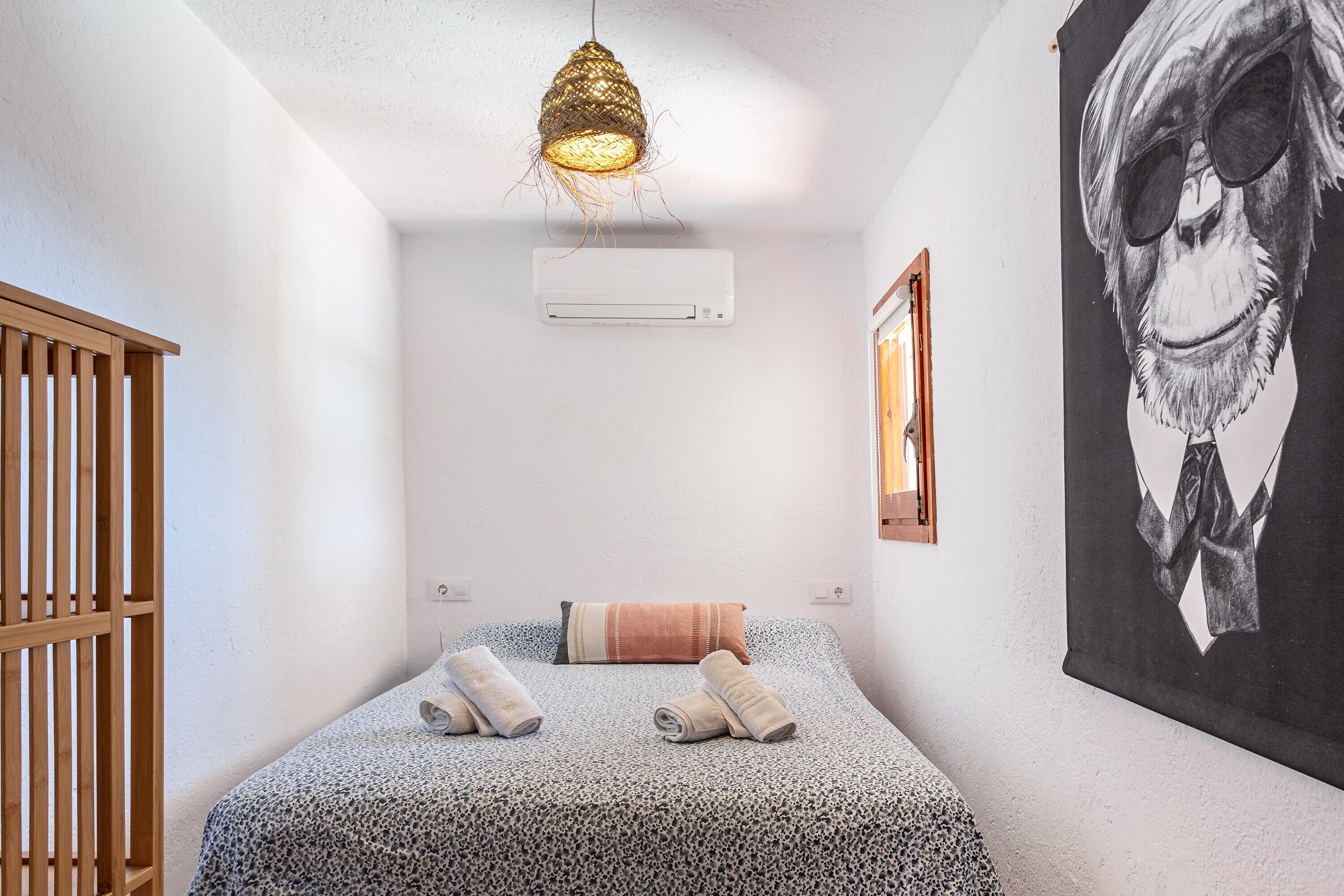 A bedroom with a bed, a hanging light fixture, an air conditioning unit, and a wall-mounted picture.