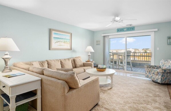 A living room with a beige sofa, a small white table, two lamps, a framed picture on the wall, and a balcony with a view.