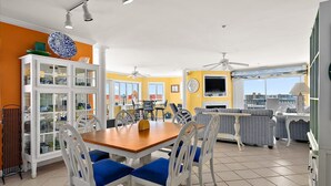 Un comedor con una mesa de madera y sillas azules, un armario blanco con puertas de cristal y un televisor montado en la pared.