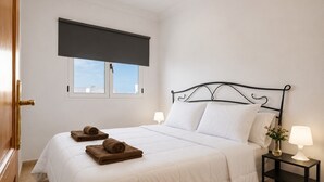 Una cama bien hecha con ropa blanca y toallas marrones, un cabecero de metal negro, una ventana con persiana oscura y una mesita de noche con una lámpara y un jarrón de flores.