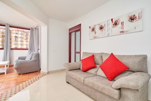A living room with a beige sofa, red cushions, a patterned rug, a white table, and a large window with curtains.