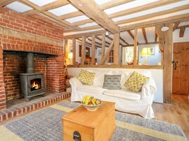 A cozy living room with a brick fireplace, wooden beams, a white sofa with patterned cushions, and a fruit bowl on a wooden table.