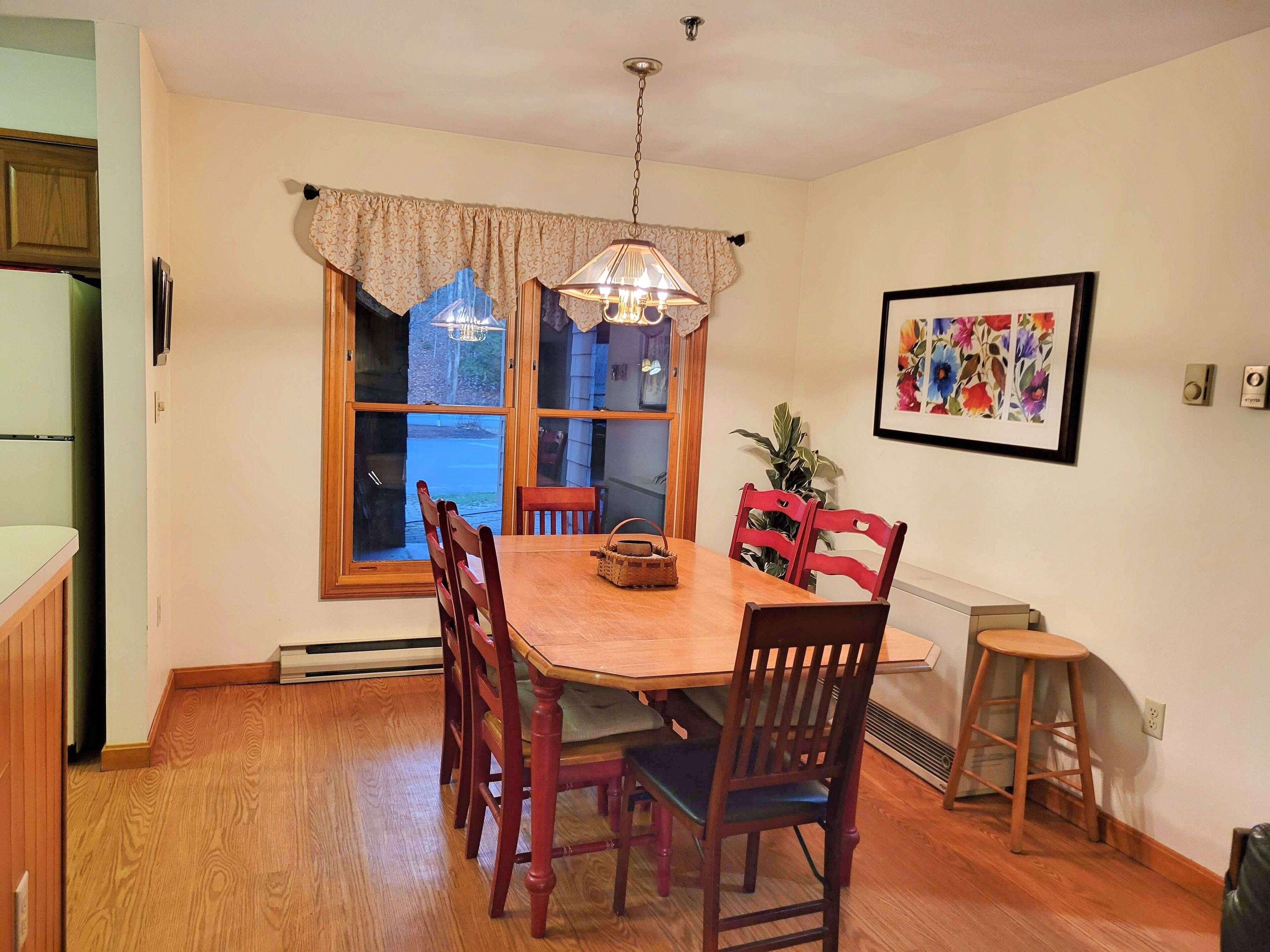 Une salle à manger avec une table en bois, six chaises, un lustre, une fenêtre avec des rideaux, un tableau floral encadré et un radiateur.
