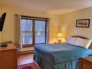 A bedroom with a bed, a desk, a lamp, a window with curtains, and a framed picture on the wall.