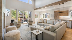 A spacious living area with a large sectional sofa, a round coffee table, and a fireplace. There are multiple windows providing natural light and a view of the outdoors. - Hummingbird House | Luxury | River | Spa (Deschutes County)