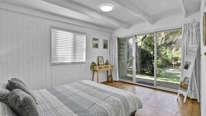 Ein Schlafzimmer mit einem großen Bett, einem Holznachttisch, einem Fenster mit Jalousien und einem Garten, der durch eine Schiebetür sichtbar ist.
