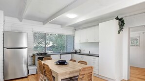Eine moderne Küche mit weißen Schränken, einem Edelstahlkühlschrank, einem Holzesstisch mit Korbstühlen und einem Fenster mit Jalousien.